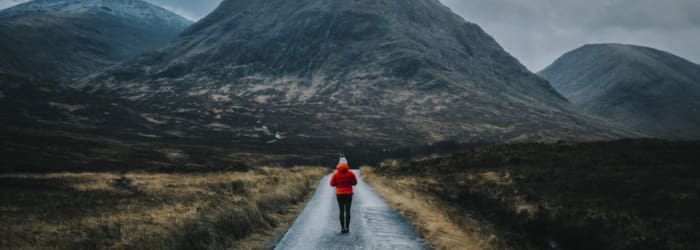 A women in a red jacket walking alone along a road in the highlands scotland with a mountain in front of her on a grey day